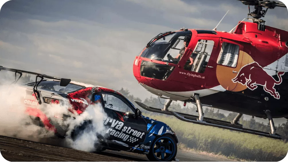 A dynamic action shot of a Red Bull sponsored helicopter flying low over a drifting race car on a dirt track, showcasing extreme sports marketing.