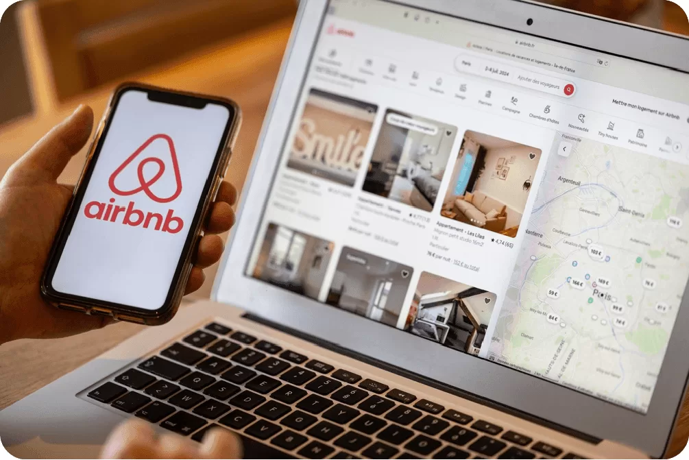 A person holding a phone with the Airbnb app open in front of a laptop displaying the Airbnb website, demonstrating a multi-platform brand presence.