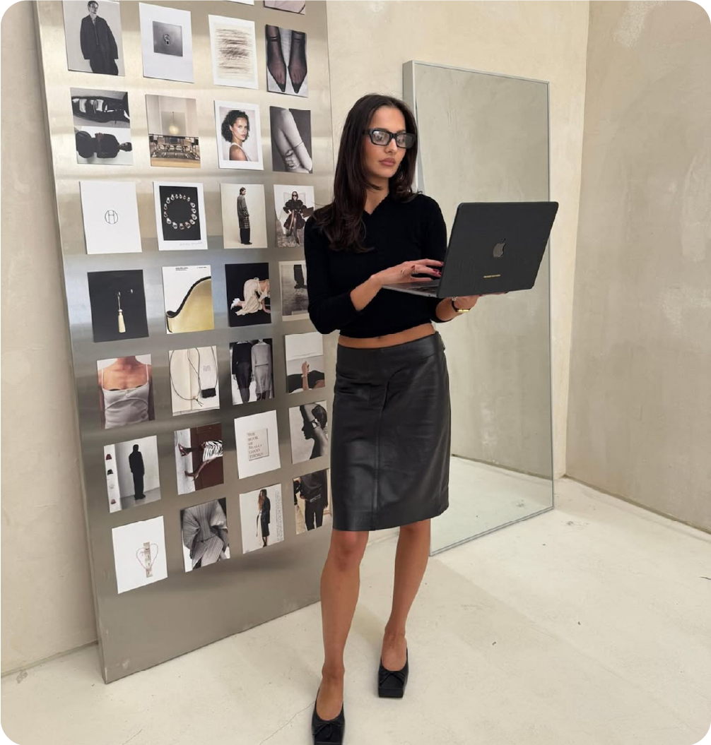 Fashion brand founder standing in a creative studio holding a laptop next to a mood board of editorial photography and design inspiration.
